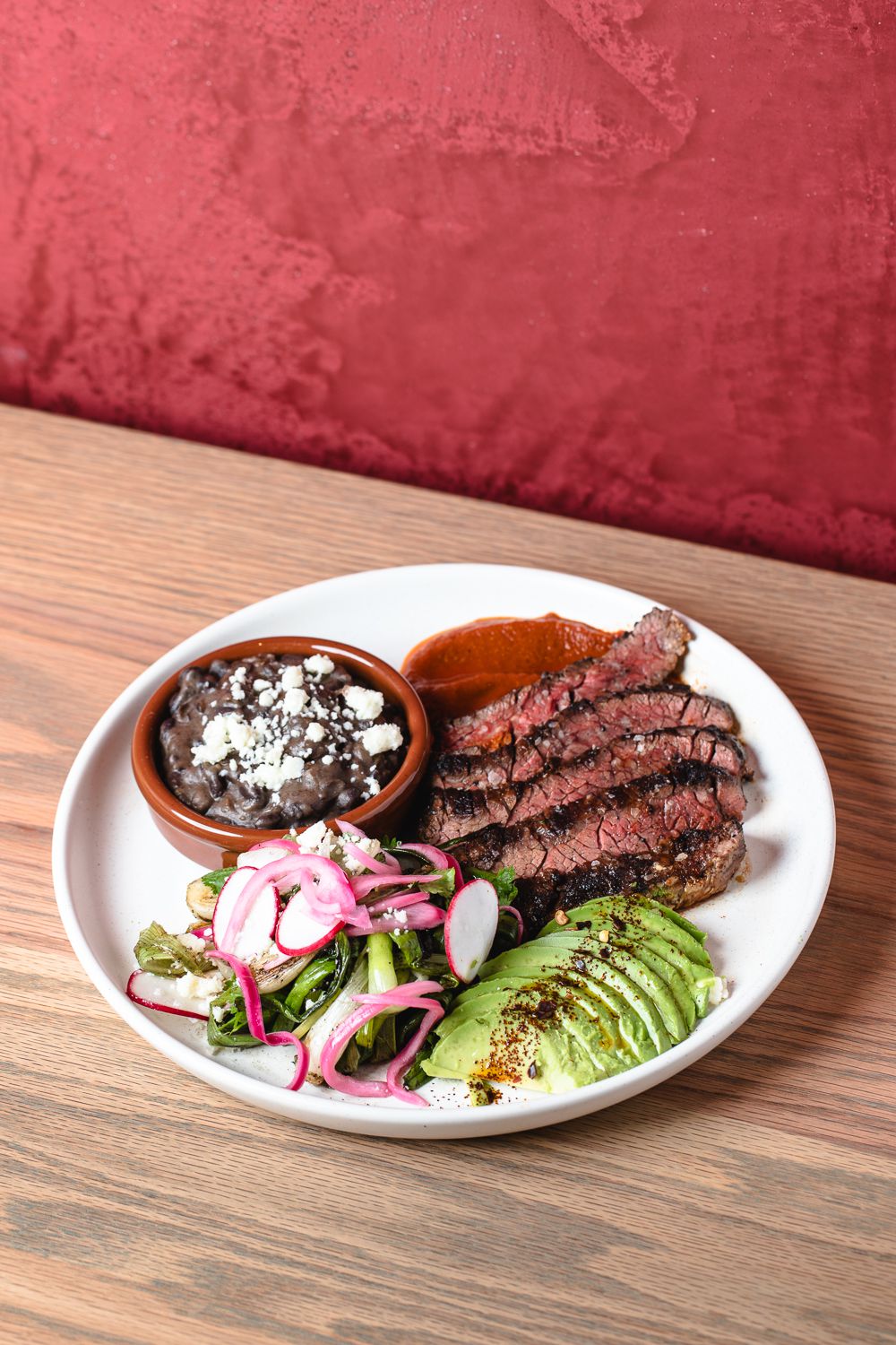 Slices of medium rare carne asada accompanied by sliced avocado, black beans, greens, pickled onions, and radishes from Chicheria MX Kitchen in Atlanta.